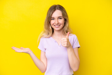 Wall Mural - Blonde English young girl isolated on yellow background holding copyspace imaginary on the palm to insert an ad and with thumbs up