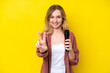 Young singer caucasian woman picking up a microphone isolated on yellow background smiling and showing victory sign