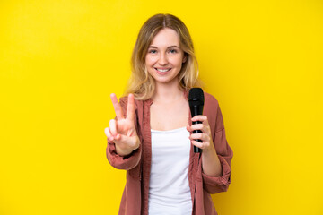 Wall Mural - Young singer caucasian woman picking up a microphone isolated on yellow background smiling and showing victory sign