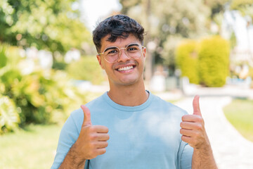 Wall Mural - Young hispanic man at outdoors With glasses and with thumb up