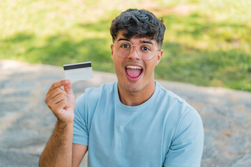 Wall Mural - Young hispanic man holding a credit card at outdoors with surprise and shocked facial expression