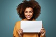 © Niko_Dali - A girl with afro curls holds a blank paper with a mockup in her hands. Person with white banner isolated on studio background.