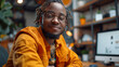 Confident young man with dreadlocks wearing glasses and a yellow jacket, smiling in a modern office environment.