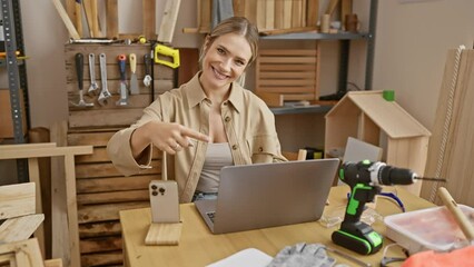 Canvas Print - Cheerful young blonde woman excitedly presenting carpentry idea, confidently pointing finger while smiling at laptop in workshop