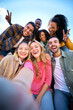 © CarlosBarquero - Vertical. Group of multiethnic friends taking selfie with smart mobile phone outdoors. Happy young multiracial people smiling together looking camera. University students having fun in college campus