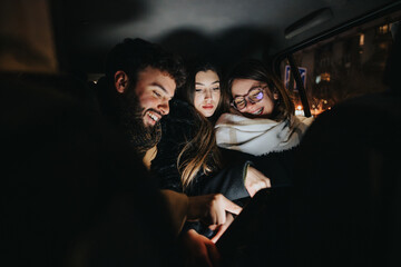 A cozy scene of three young adults gathered around a smart phone, sharing a moment of connection in a low-lit indoor setting at night.