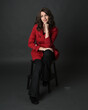 full length portrait of beautiful brunette woman model, wearing red trench coat jacket with black leather pants. sitting pose with hand gesture on dark grey studio background.