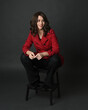 full length portrait of beautiful brunette woman model, wearing red trench coat jacket with black leather pants. sitting pose with hand gesture on dark grey studio background.