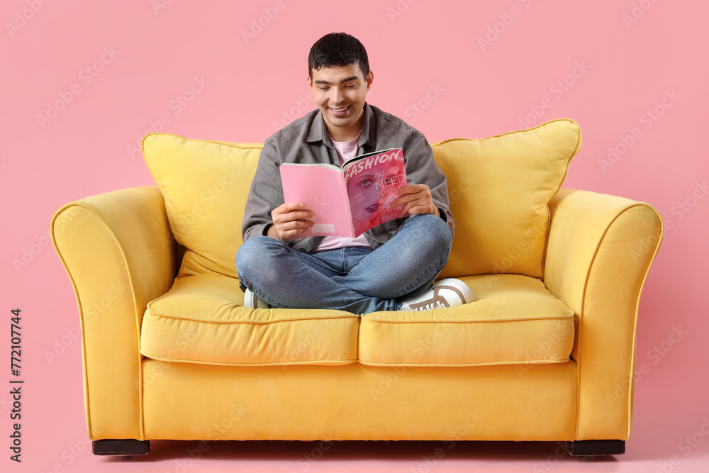 Handsome young man reading magazine on yellow sofa against pink background