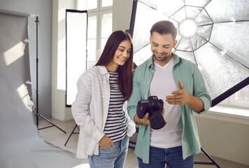 Portrait of a young smiling professional photographer showing photos to a girl model on digital camera standing in production studio with light equipment and discussing photo session.