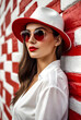 Portrait stylish young woman model in sunglasses standing at wall wearing white shirt and red hat, looking away. Fashionable lady posing outdoors, lifestyle. Fashion style concept. Copy text space