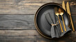 Black plate with gray textile napkin and brass flatware on a light wooden surface