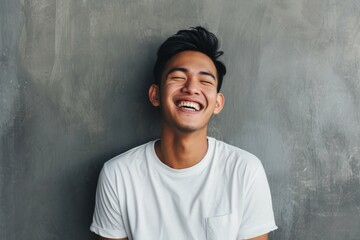 Wall Mural - Joyful young man laughing with eyes closed, leaning against a gray background, wearing a white t-shirt, embodying happiness and positivity.