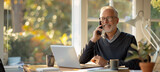 Fototapeta  - Senior Businessman on Call with Sunny Office Background. Mature businessman in a smart sweater having a pleasant conversation on the phone in a bright office with a lush green backdrop.