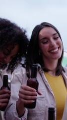 Wall Mural - Vertical. Cheerful multiracial young people toast with beer bottles celebrating dancing and laughing on summer outdoors. Group happy millennial friends together enjoying sunset drinks at rooftop party