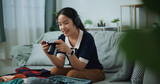 Fototapeta Mapy - Selective focus of Asian teenager woman wear wireless headphones sitting on sofa look pictures on the camera while packing suitcase luggage in the living room , Preparation travel suitcase at home.