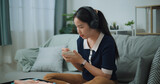 Fototapeta Mapy - Selective focus of Asian teenager woman wear wireless headphones sitting on sofa making checklist of things to pack for travel, Preparation travel suitcase at home.