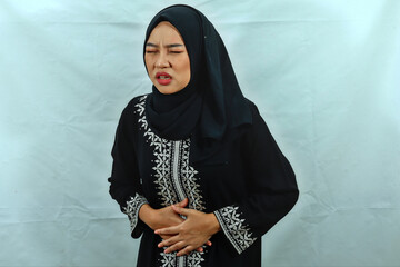 Unhealthy beautiful young Asian Muslim woman wearing hijab and black dress with white pattern touching belly, having stomach pain isolated on white studio background
