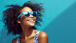 Fashion portrait of a young, stylish, happy and smiling African American woman with sunglasses on bright blue background.