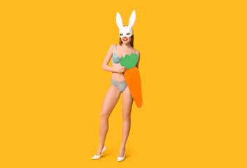 Poster - Sexy smiling young woman in bunny mask with paper carrot on yellow background. Easter celebration