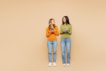 Full body young friends two women wearing orange green shirt casual clothes together hold in hand use mobile cell phone look to each other isolated on plain pastel beige background. Lifestyle concept.