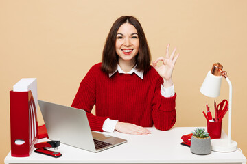 Wall Mural - Young smiling happy employee business woman wear red sweater shirt sit work at office desk with pc laptop showing okay ok gesture isolated on plain pastel beige background. Achievement career concept.