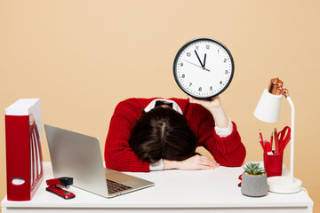 Wall Mural - Young sad employee business woman wear red sweater sit work at office desk with pc laptop hold in hands clock put head on table isolated on plain pastel beige background. Achievement career concept.