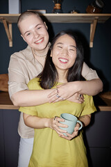 Vertical medium flash portrait of cheerful young adult Caucasian woman standing in kitchen hugging her Asian girlfriend and smiling at camera