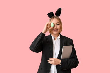 Wall Mural - Happy adult businesswoman in bunny ears with tablet computer and egg on pink background. Easter celebration