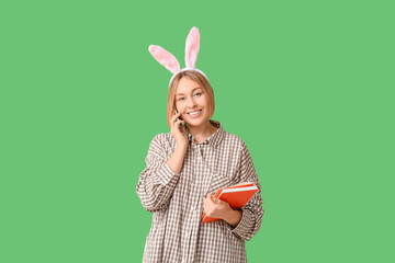 Wall Mural - Happy adult female manager in bunny ears with notebook talking by mobile phone on green background. Easter celebration