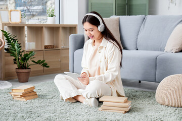 Wall Mural - Beautiful young Asian woman in headphones listening to audiobook with books and modern tablet computer on carpet in living room