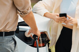 Fototapeta Zachód słońca - Young couple use smartphone to pay for electricity at public EV car charging station green city park. Modern environmental and sustainable urban lifestyle with EV vehicle. Expedient