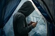 individual with hoodie using a smartphone in the opening of their tent