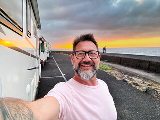 One happy man taking selfie picture on the side of a modern camper van motorhome. Traveler vanlife lifestyle people sharing on social media adventure and journey. Amazing sunset in background. Parking