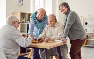 Group of happy diverse senior people playing chess in nursing home sitting at the table. Pensioners spending leisure time together playing board games. Leisure in retirement home concept.