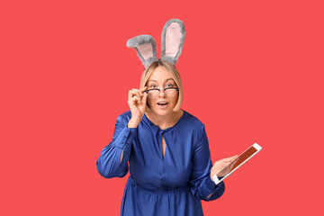 Wall Mural - Surprised adult businesswoman in bunny ears with tablet computer on red background. Easter celebration