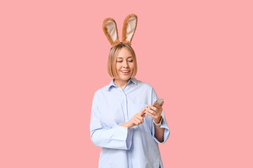 Wall Mural - Happy adult businesswoman in bunny ears with mobile phone on pink background. Easter celebration