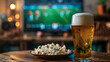 A glass of beer and popcorn stand on a table in a sports bar against the backdrop of a TV showing football