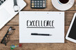 Excellence written in notepad on office desk with computer technology, high angle