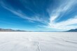 A vast salt flat with cracked white surface stretches to mountains under a sweeping blue sky with wispy clouds.