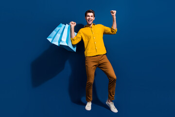 Sticker - Full size photo of handsome young man raise fists shopping bags promo wear trendy yellow outfit isolated on dark blue color background