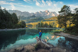 Fototapeta  - Man enjoying the amazing morning scenery at a gorgeous lake in the Bavarian Alps, with teal water reflecting the view of the mountain range and the nice clouds