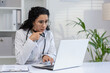 © Liubomir - Latino woman doctor using laptop, looking puzzled or concerned, in a medical office setting.