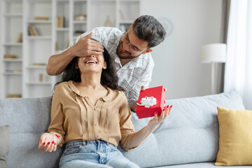 Loving indian man covering woman eyes and giving present gift box, guy making surprise for his wife on birthday, home interior