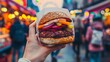 Hand holding a vegan burger with a colorful patty and fresh vegetables, with the lively ambiance of a street food market in the background.