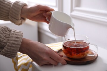 Wall Mural - Woman pouring milk into cup of tea at white table, closeup