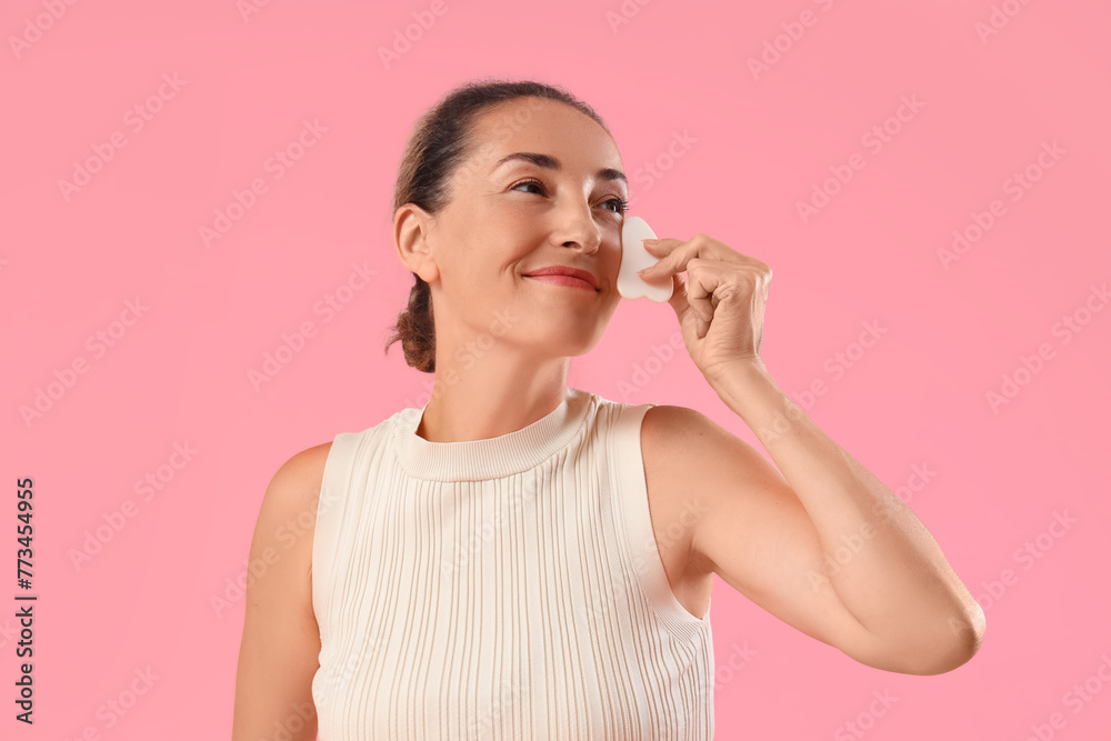 Beautiful mature woman using guasha for face massage on pink background