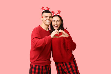 Wall Mural - Happy young couple in Christmas pajamas making heart with their hands on pink background