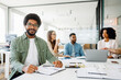 © Vadim Pastuh - A confident man in a casual green shirt and glasses smiles at the camera, surrounded by focused colleagues in a modern office setting, suggesting a collaborative and inclusive work environment