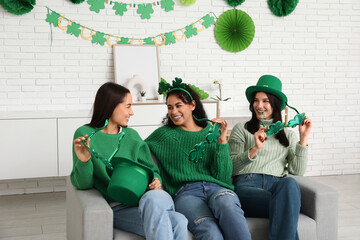 Sticker - Beautiful young women with decorative glasses in shape of clover on sofa at home. St. Patrick's Day celebration